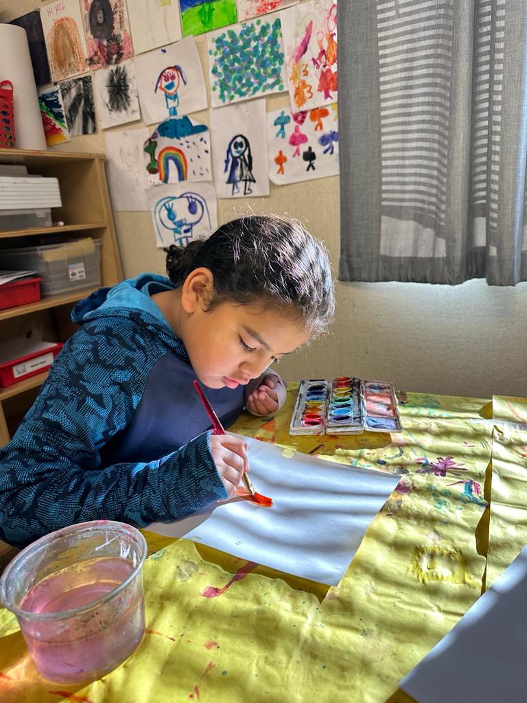 kid painting using red paint at a table with art in the background.