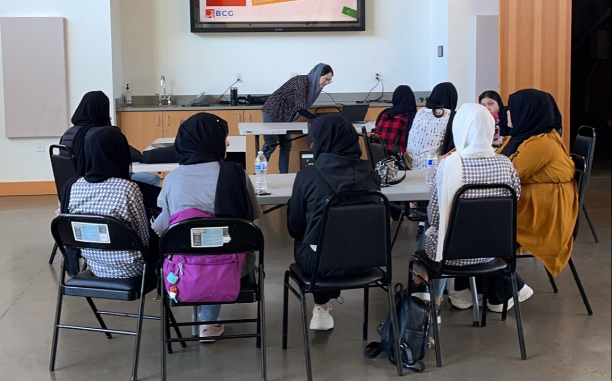 Students work on laptops while sitting around tables together with an instructor